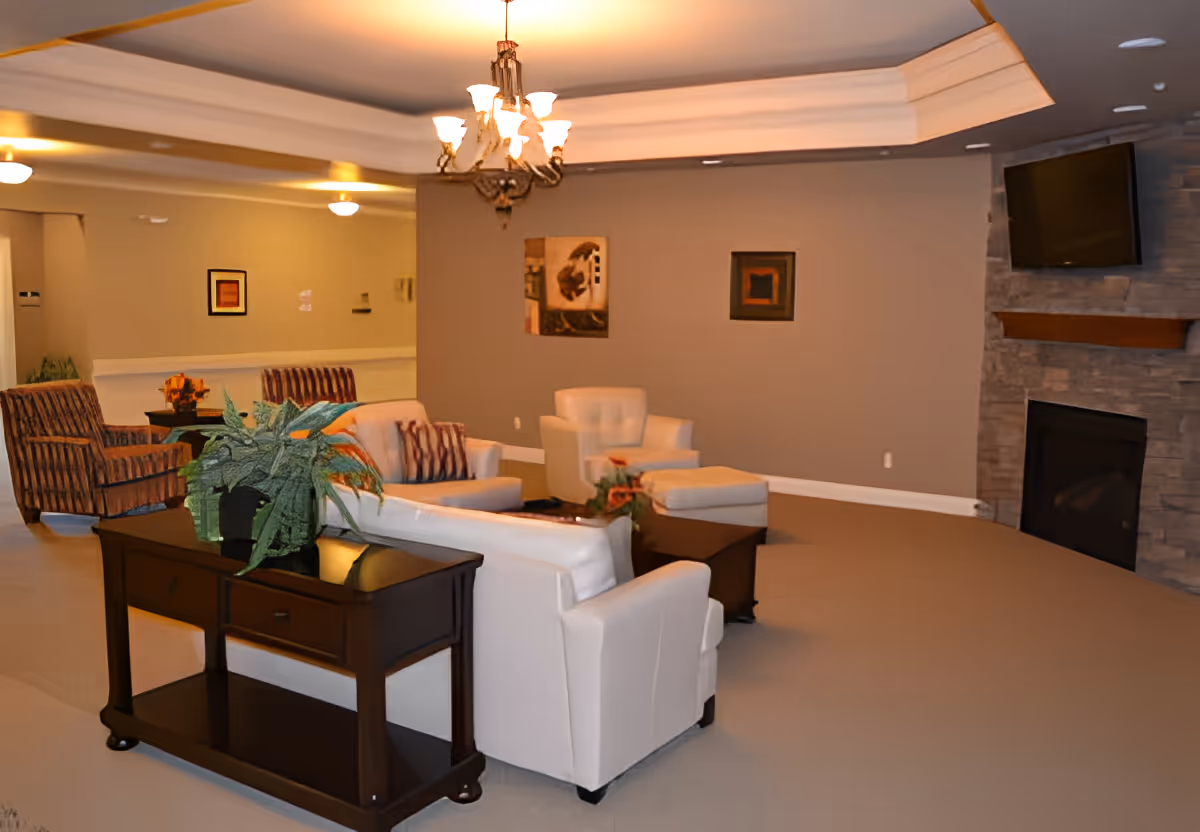 A cozy living room area in Autumn Fields Adult Community-Hoopeston featuring white and striped armchairs, a dark wooden console table with green plants, a stone fireplace with a mounted flat-screen TV above it, and warm lighting from a chandelier and ceiling lights. The walls are decorated with framed artwork.