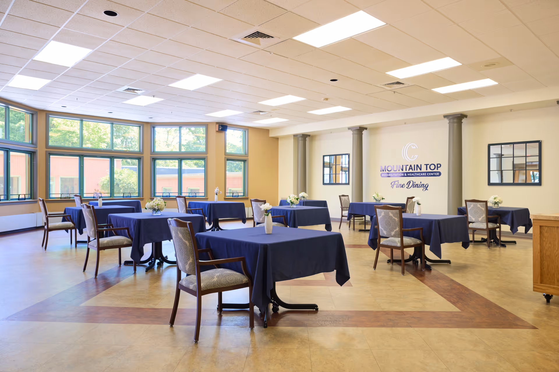 A spacious dining room with multiple tables covered in dark blue tablecloths, each table adorned with a small vase of flowers. The room has large windows letting in natural light, beige walls with two mirrors, and a sign on the wall that reads 'Mountain Top Rehabilitation & Healthcare Center Fine Dining'.