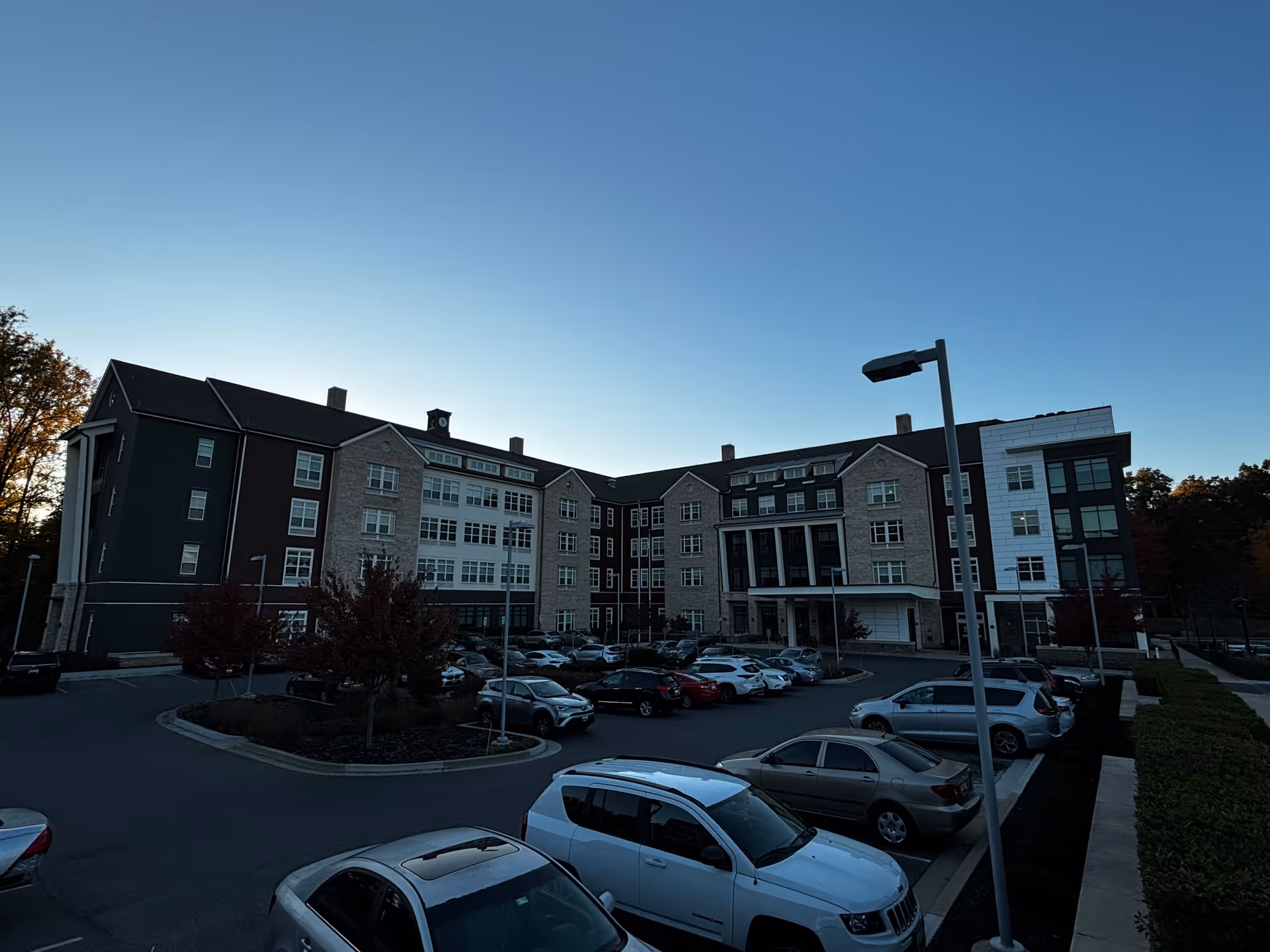 Front view of a multi-story senior living building with a parking lot and parked cars under a clear evening sky.