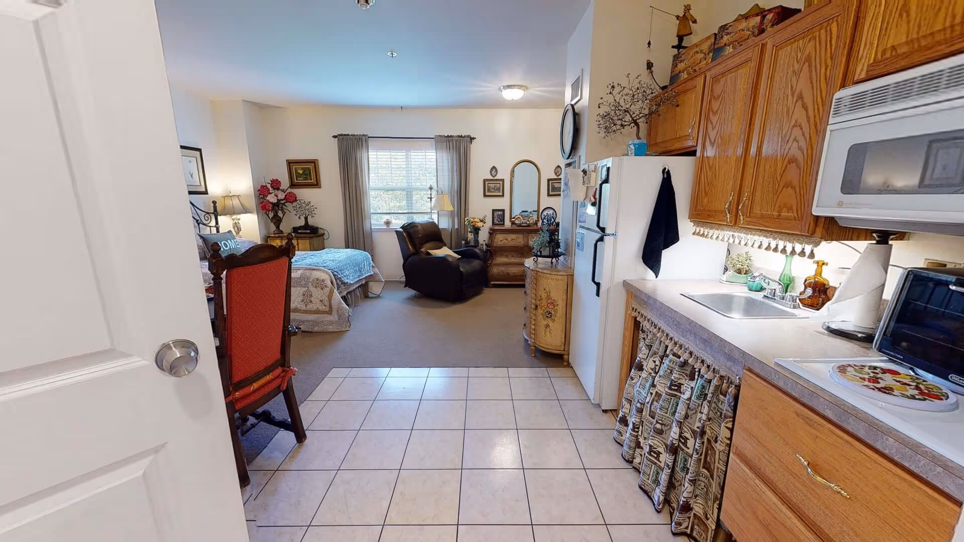 View of a cozy assisted living apartment showing a small kitchen area with wooden cabinets, a microwave, a refrigerator, and a sink on the right side. The kitchen floor is tiled. Beyond the kitchen is a living and sleeping area with a bed, an armchair, a window with curtains, and various decorative items including framed pictures and flowers.
