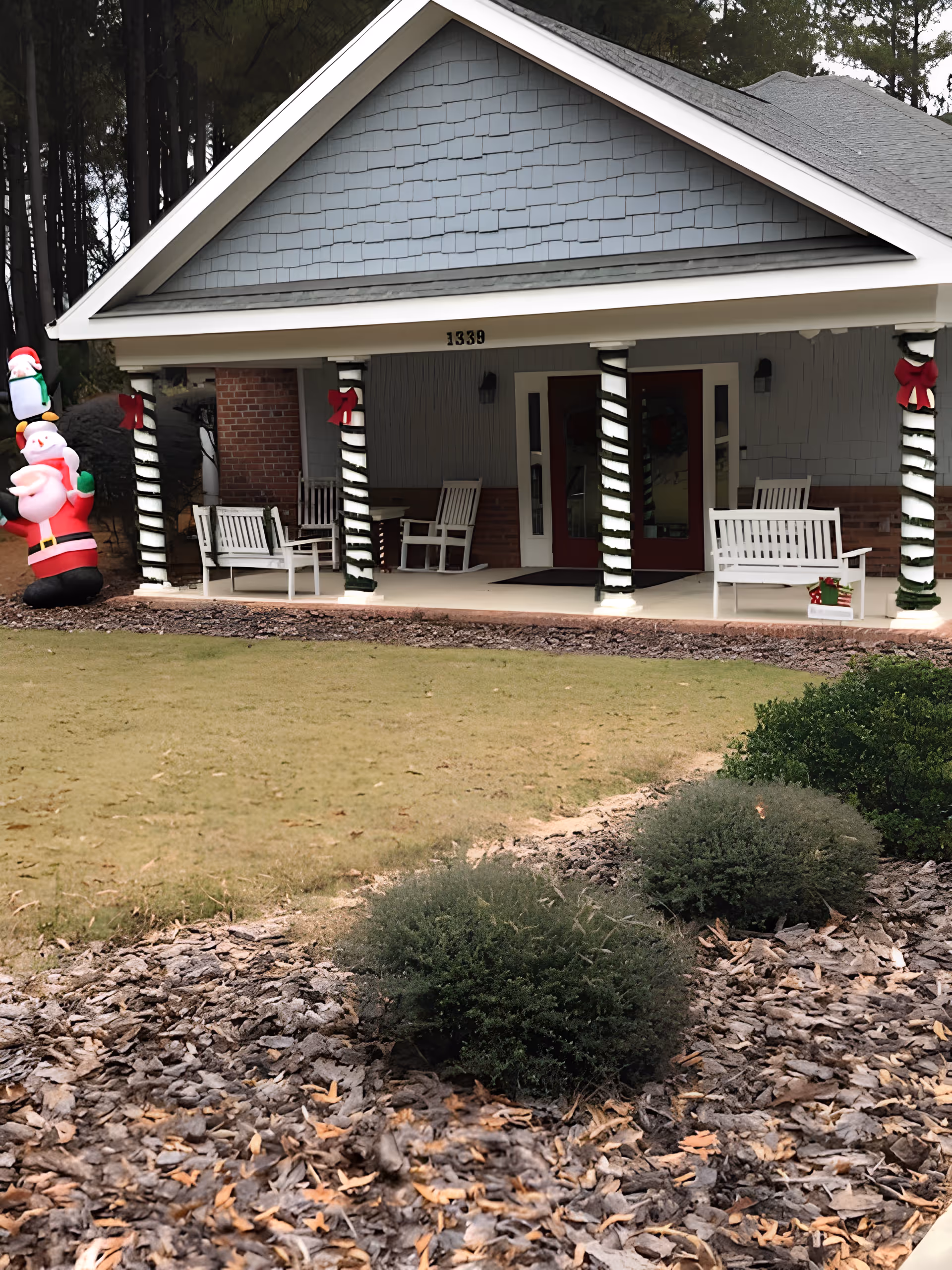A front porch entry with white rocking chairs, columns wrapped in garland and red bows, and an inflatable Santa on the lawn.
