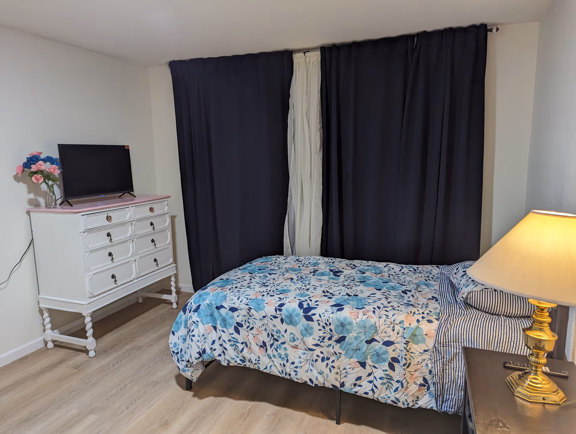 Small bedroom with a single bed in floral bedding, a dresser with a TV and flowers, dark curtains, and a bedside lamp.