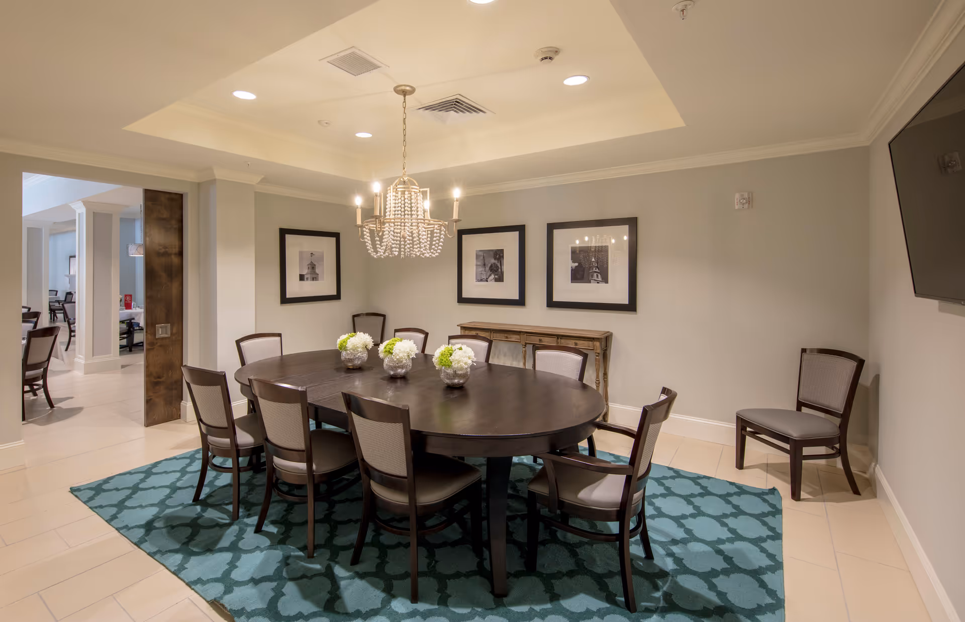 Round dark wood dining table with eight chairs on a patterned teal rug under a crystal chandelier in a bright dining room with framed artwork on the wall.