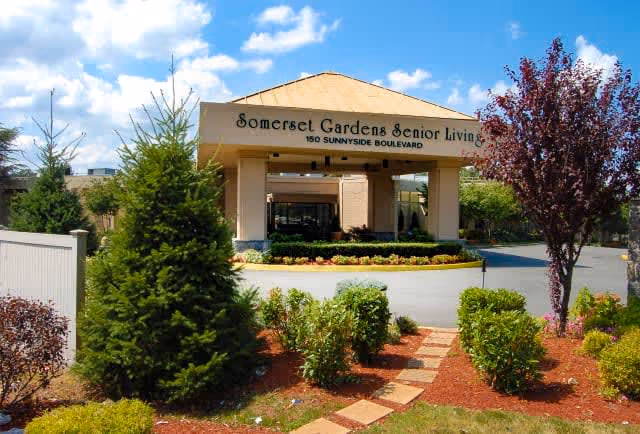 Entrance to Somerset Gardens Senior Living facility with a covered driveway and landscaped garden featuring bushes, small trees, and a stone pathway under a blue sky with some clouds.
