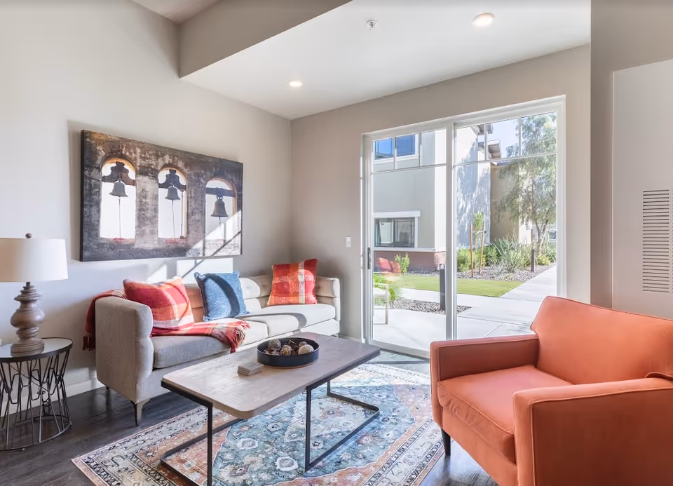 A bright living room with a gray sofa adorned with red and blue pillows, an orange armchair, a wooden coffee table on a patterned rug, a side table with a lamp, and a large window showing an outdoor garden area.