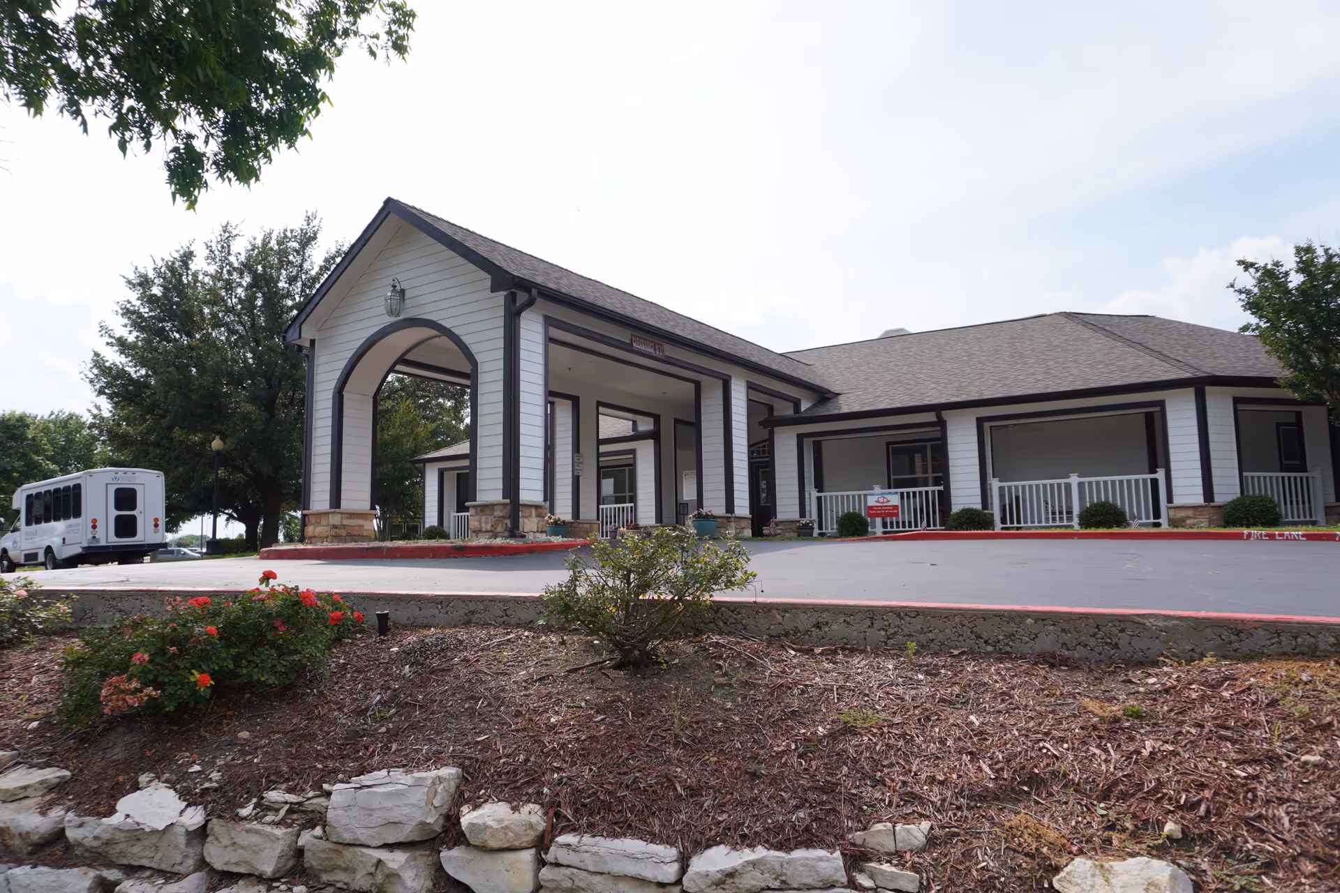 Exterior view of a single-story building with white siding and a covered entrance supported by columns. There is a small landscaped area with bushes and flowers in front, and a white shuttle bus parked on the left side. The sky is partly cloudy.
