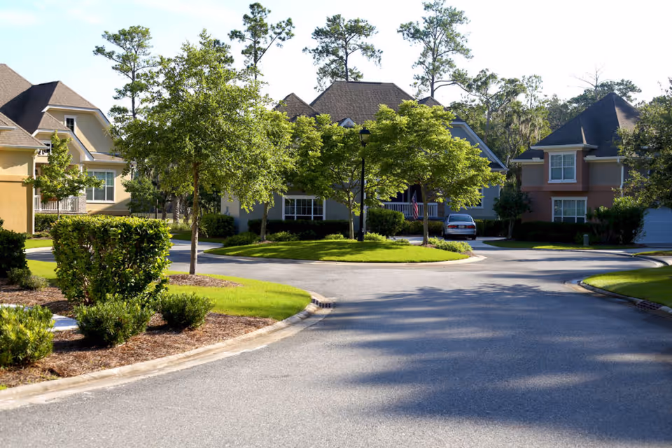 A quiet residential street in a senior living community with well-maintained houses, green lawns, trees, and shrubs under a clear sky.