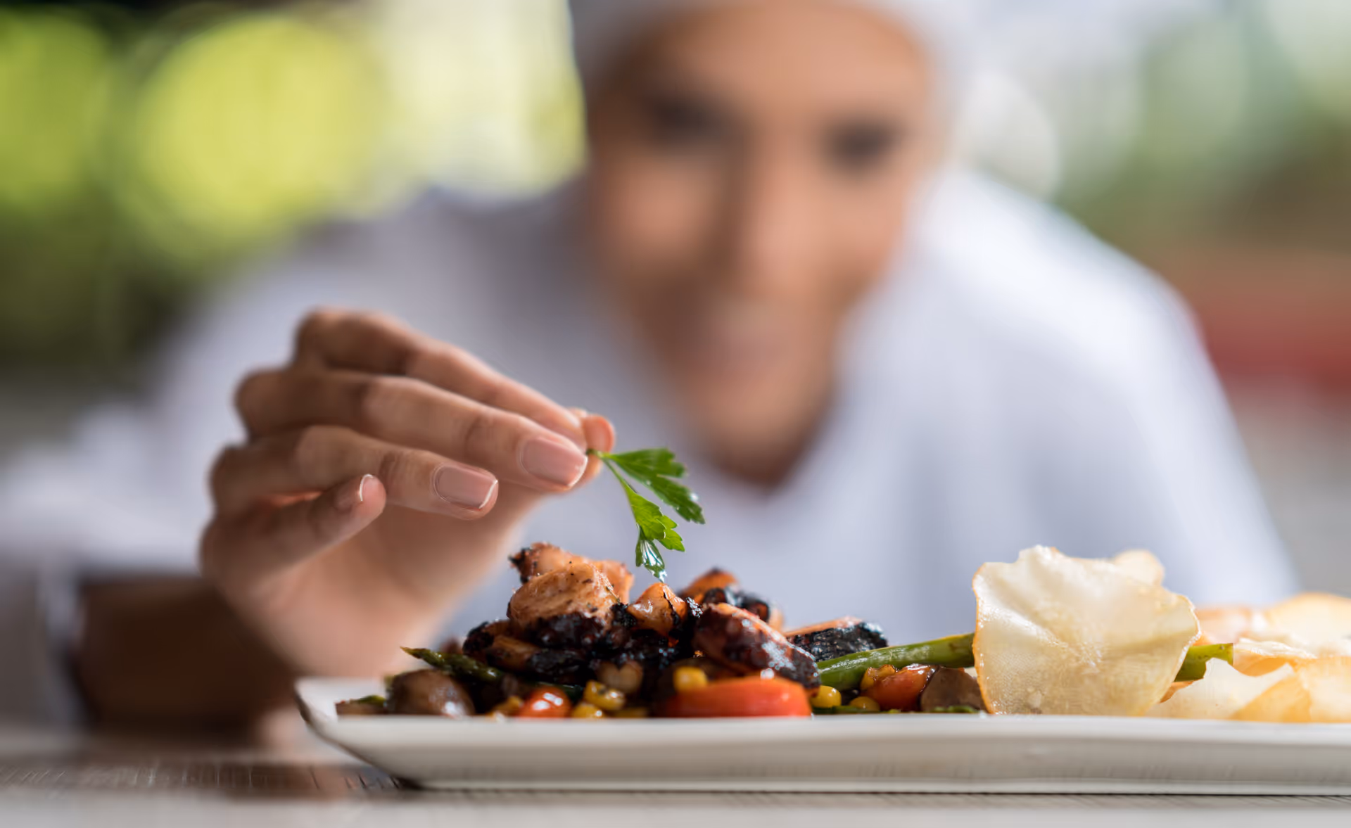 A chef carefully garnishing a plated dish with a sprig of parsley, with the chef's face blurred in the background.