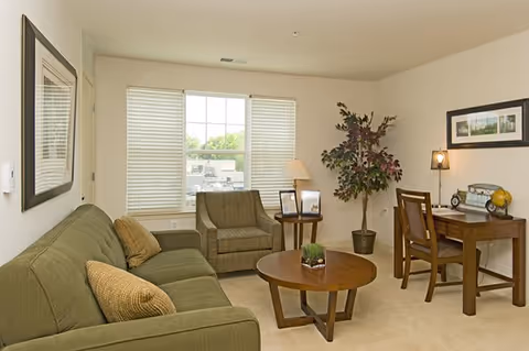 A cozy living room with a green sofa, two beige cushions, a wooden coffee table with a small plant, an armchair next to a window with white blinds, a side table with a lamp and picture frames, a potted plant, and a wooden desk with a chair, lamp, and decorative items.
