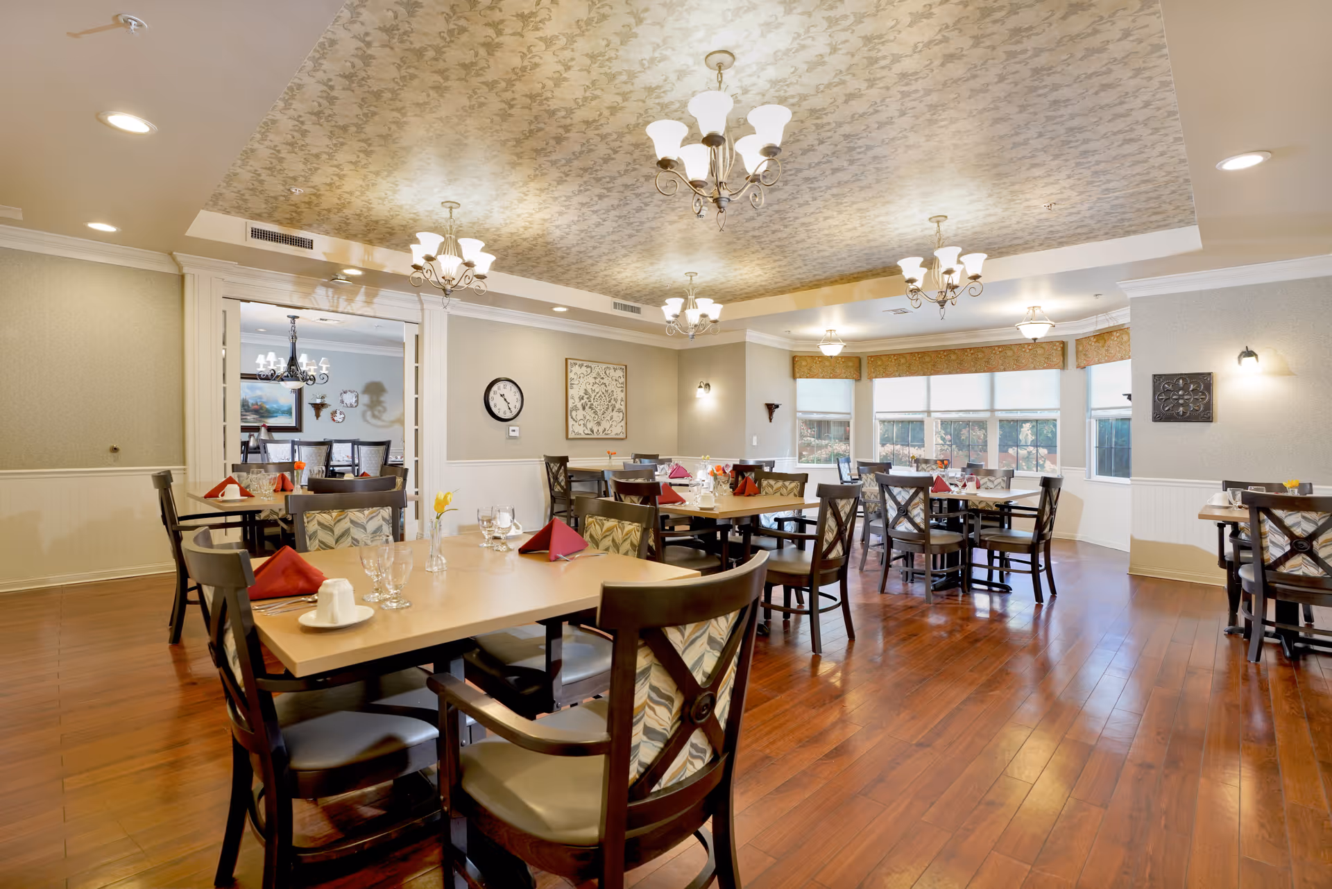 A spacious dining room with wooden floors, multiple tables set with glasses, cups, and red napkins, surrounded by wooden chairs with patterned cushions. The ceiling has decorative wallpaper and multiple chandeliers providing warm lighting. Large windows with blinds allow natural light to enter the room.