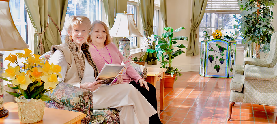 Two elderly women sit on a floral couch in a sunlit lounge with plants, lamps, and large windows.