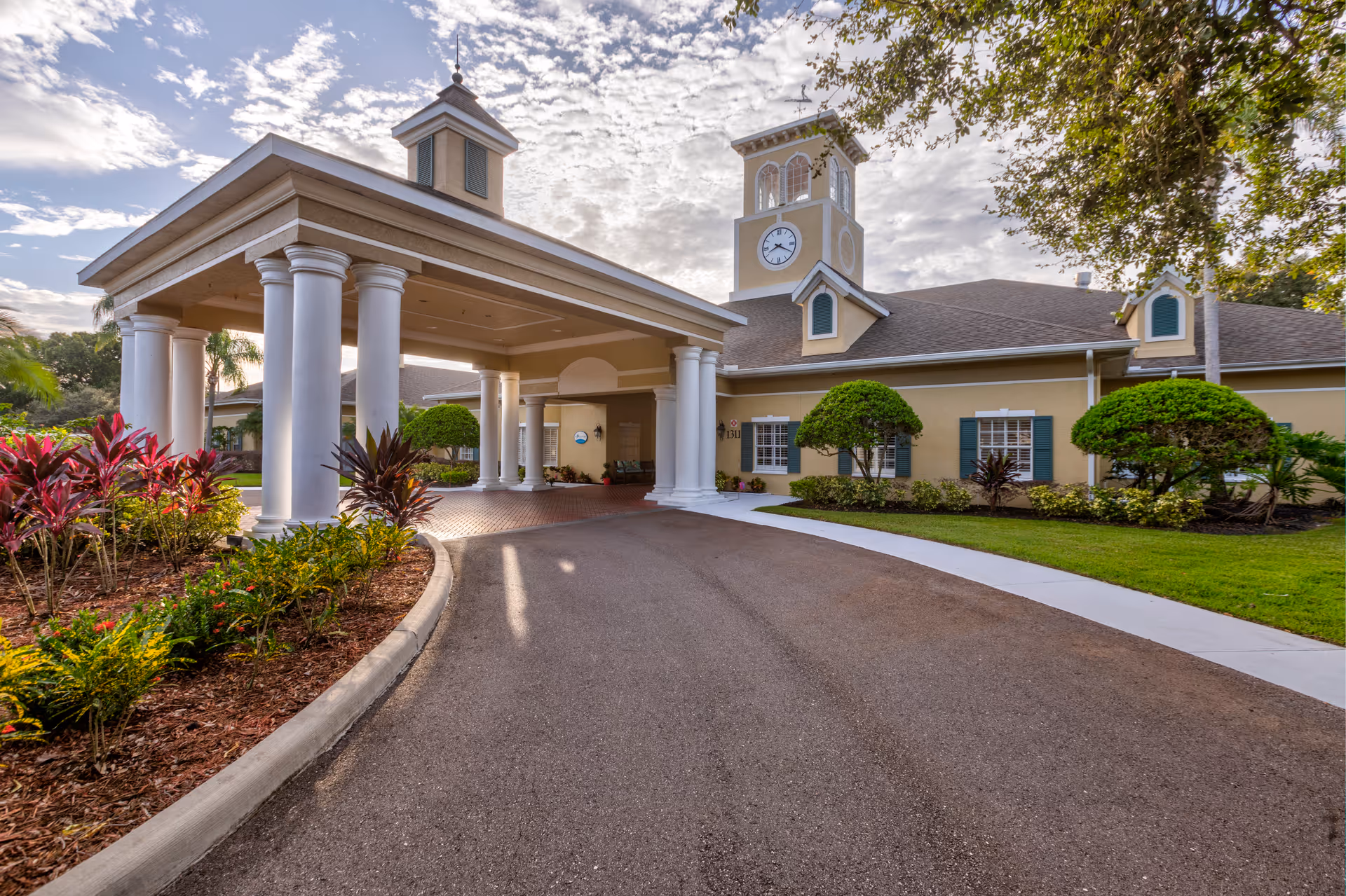 Exterior view of a senior living facility building with a covered entrance supported by white columns, a clock tower, manicured bushes, and a curved driveway under a partly cloudy sky.