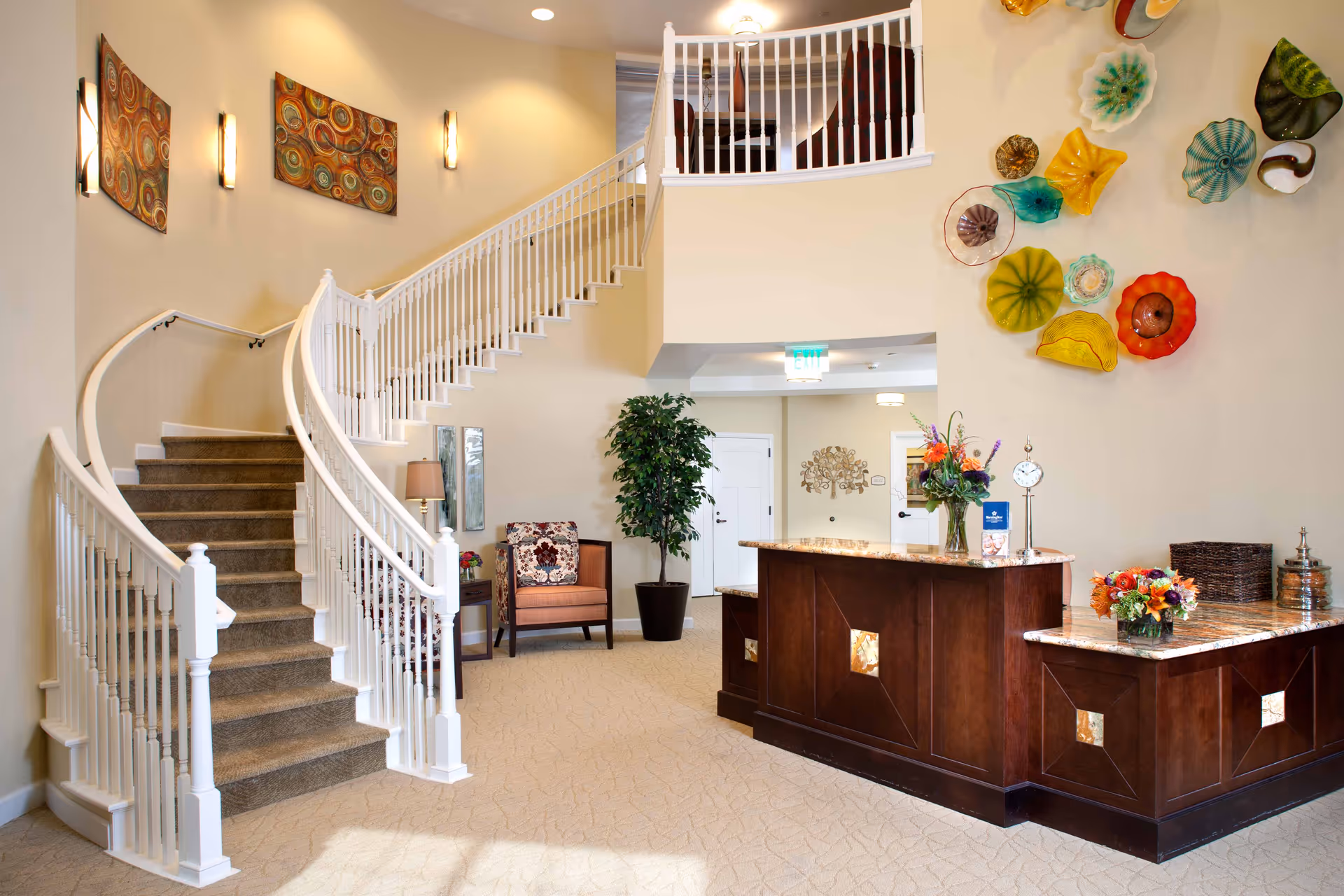 Bright and welcoming interior of a senior living facility featuring a curved staircase with white railings, a dark wood reception desk adorned with flower arrangements, a potted plant, and colorful decorative glass wall art. The space is well-lit with wall sconces and ceiling lights, and there is a comfortable chair with a floral cushion near the staircase.