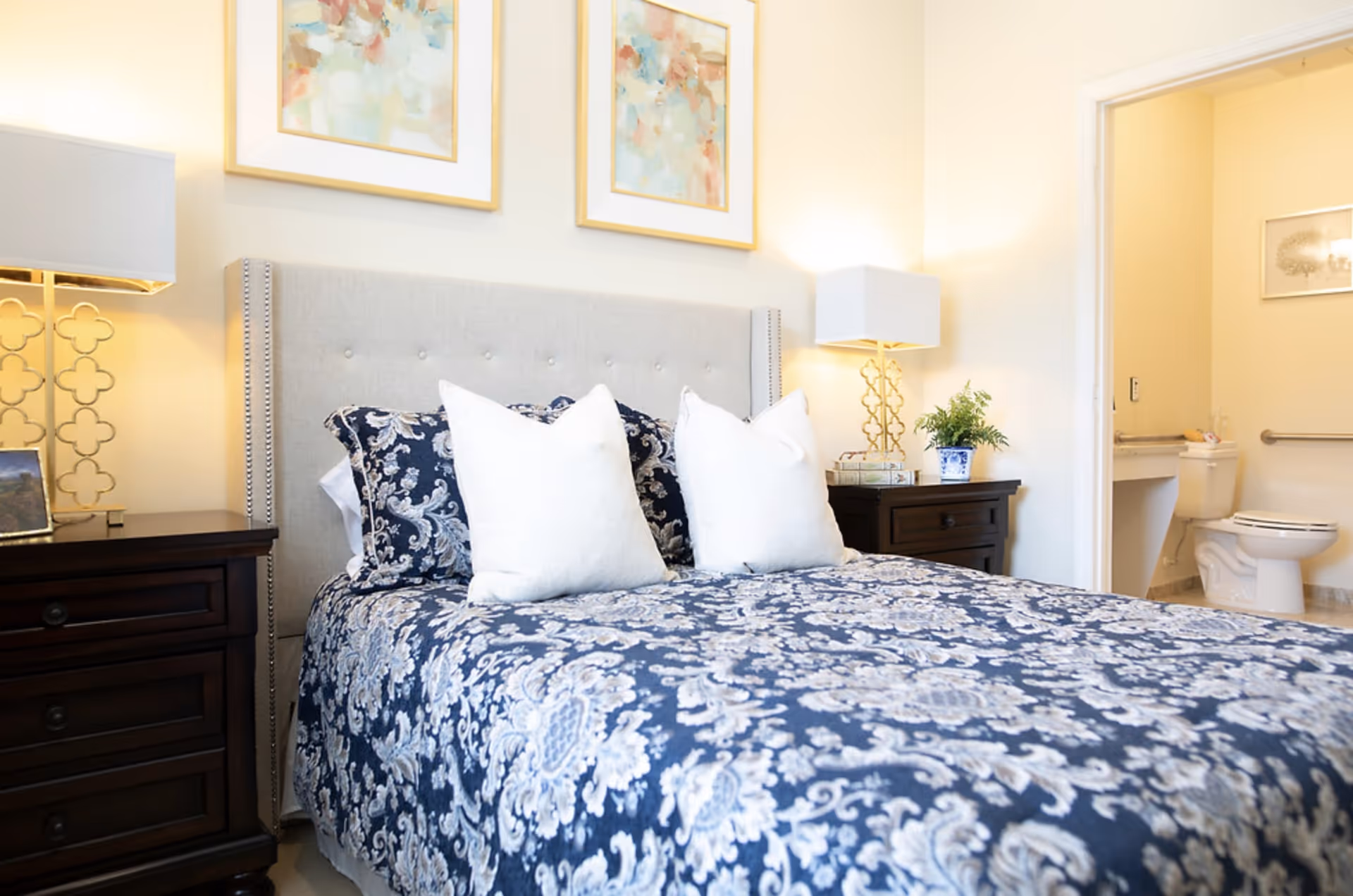 Bright bedroom with a patterned blue bedspread, two white pillows, matching nightstands and lamps, and an open doorway revealing a bathroom.