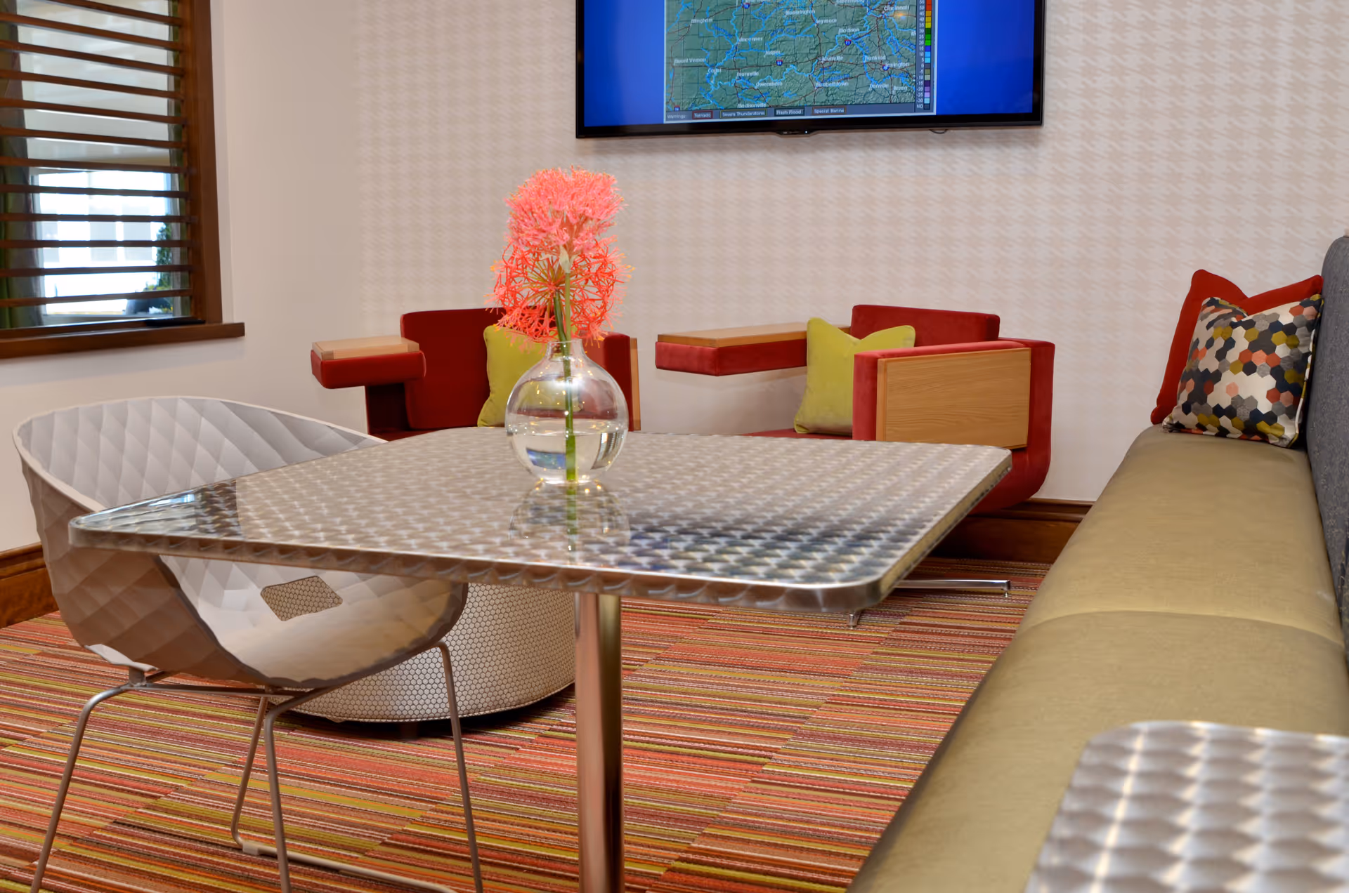 Modern lounge seating area with a metal table, contemporary chairs and sofa, colorful pillows, and a vase with pink flowers beneath a wall-mounted TV.