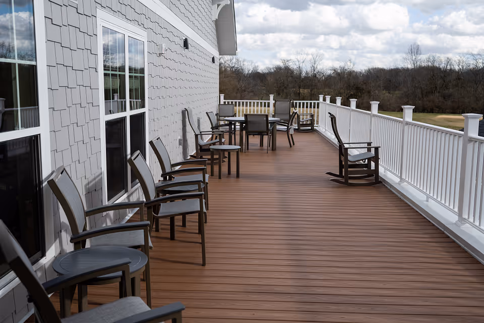 Outdoor wooden deck attached to a building with gray siding, featuring multiple chairs, tables, and rocking chairs arranged along the deck. White railing borders the deck, and trees and a cloudy sky are visible in the background.