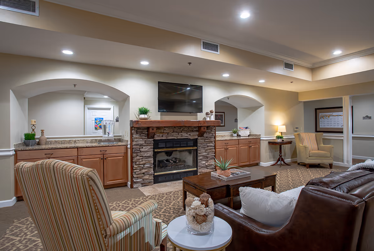 Cozy senior living common area with a stone fireplace and wall-mounted TV, surrounded by sofas, armchairs, a coffee table, and built-in cabinets.