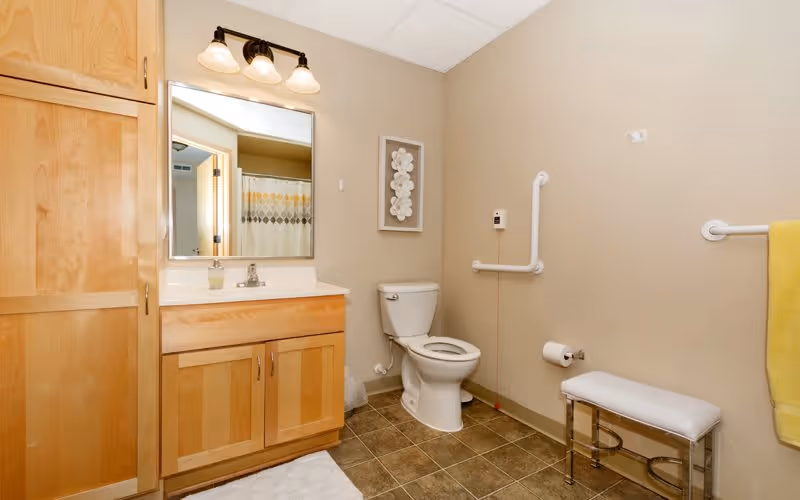 A clean and accessible bathroom featuring a wooden vanity with a sink and mirror above it, a toilet with grab bars on the wall for support, a small padded bench, a towel rack with a yellow towel, and beige walls and tiled floor.