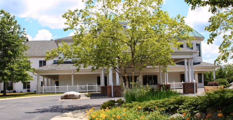 Front entrance of a light-colored senior living building with a covered porch, columns, trees, and landscaped driveway.
