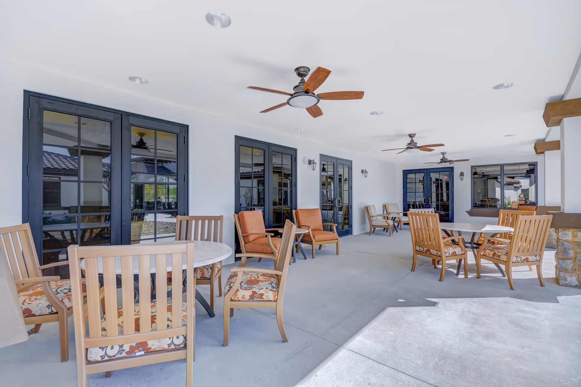 Covered outdoor patio area with multiple seating arrangements including round tables with wooden chairs featuring floral cushions and armchairs with orange cushions. The patio has ceiling fans and is adjacent to a building with large glass doors and windows.