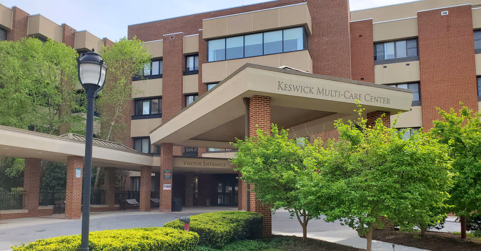 Front entrance of the Keswick Multi-Care Center, a brick multi-story building with a covered porte-cochere and landscaped shrubs.