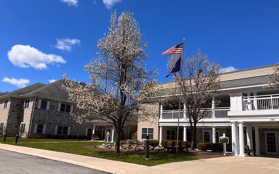 Exterior view of Westminster Woods senior living facility on a sunny day with a clear blue sky. The building features a stone and beige siding facade with white columns and railings. Two trees with white blossoms are in front of the building, along with two flagpoles flying the American flag and another flag. There is a paved driveway and green lawn surrounding the entrance.