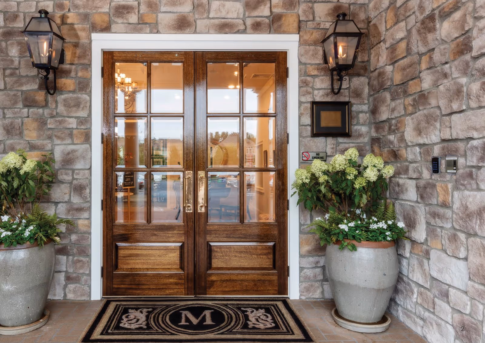 Entrance to a building with double wooden doors featuring glass panes. The exterior walls are made of stone, and there are two large planters with green and white flowering plants on either side of the doors. Two black wall-mounted lanterns are positioned above the planters. A black doormat with a decorative letter 'M' is placed in front of the doors. A small 'No Smoking' sign is visible on the right side wall.