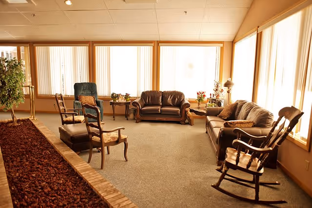 A cozy living room area with large windows covered by vertical blinds allowing natural light to fill the space. The room features two leather sofas, a blue armchair, a rocking chair, two wooden chairs, and a matching ottoman. There are small tables with potted plants and flowers, and a floor lamp near one of the sofas. A planter with a small tree and mulch runs along one side of the room.