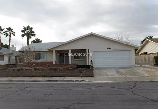 Single-story residential building with a front porch, two-car garage, and a low brick wall bordering the front yard. There are palm trees visible in the background under a cloudy sky.