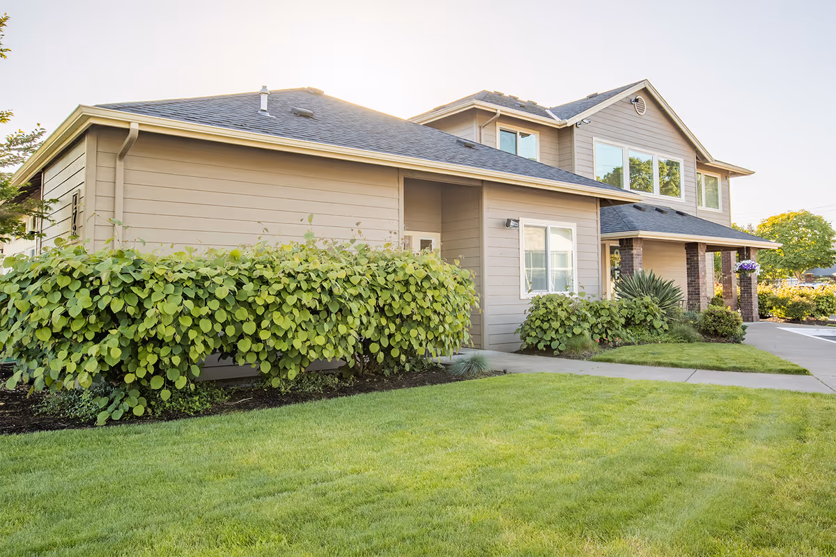 Two-story beige building with a manicured lawn, shrubs, and a covered entrance under a clear sky.