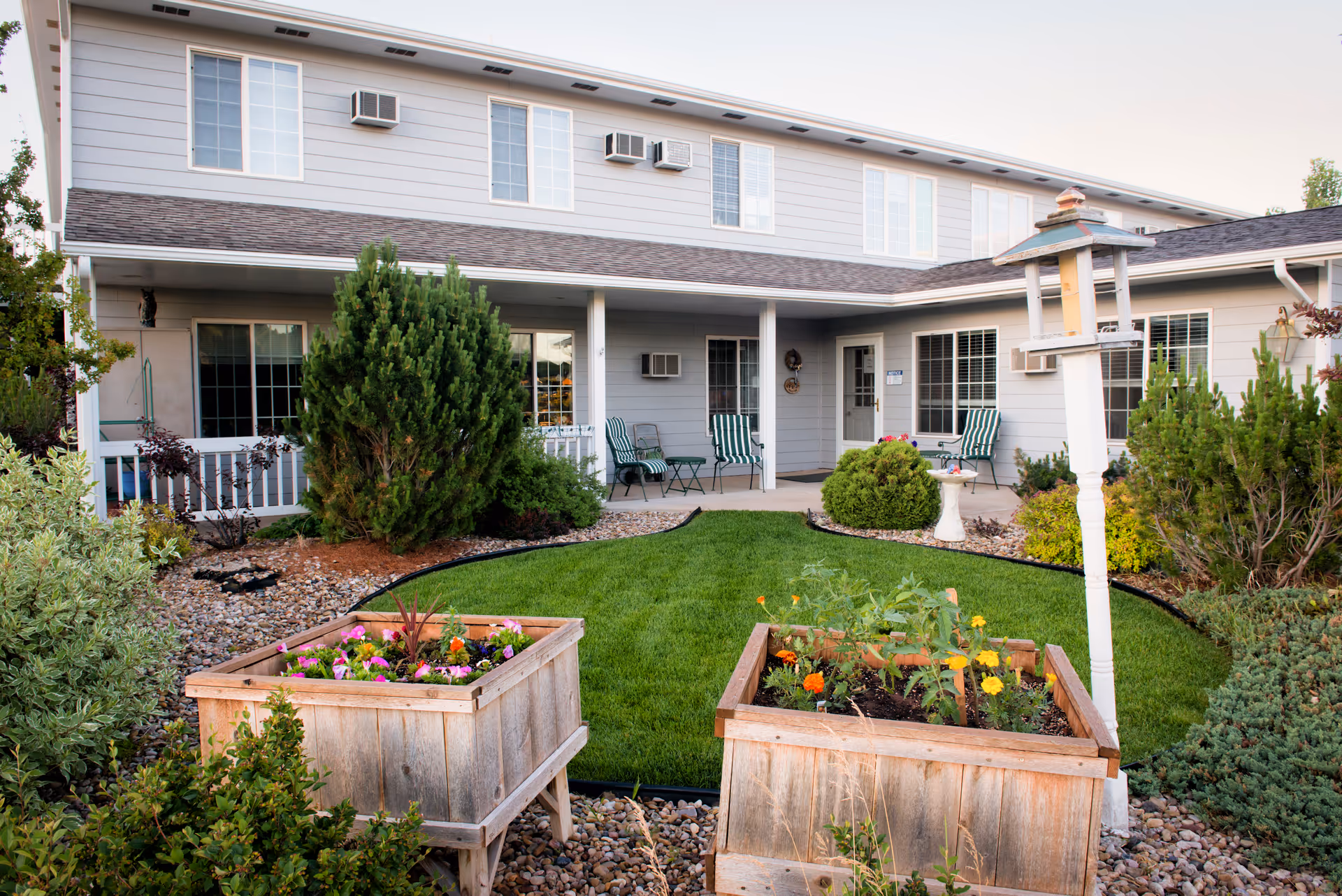 Outdoor courtyard area of a senior living facility with a well-maintained lawn, two wooden planter boxes filled with flowers, various shrubs, and a white birdhouse on a post. The building in the background has light gray siding, multiple windows, and a covered porch with green-striped chairs and small tables.
