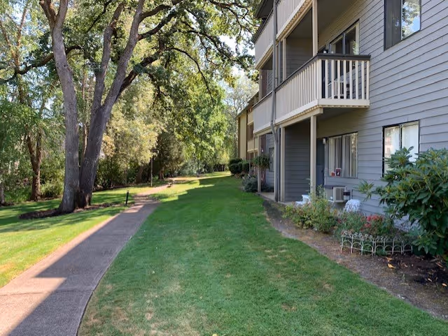 Outdoor view of a retirement residence showing a paved walkway alongside a grassy area with large trees and shrubs. The right side features a multi-story building with balconies and windows overlooking the green space.