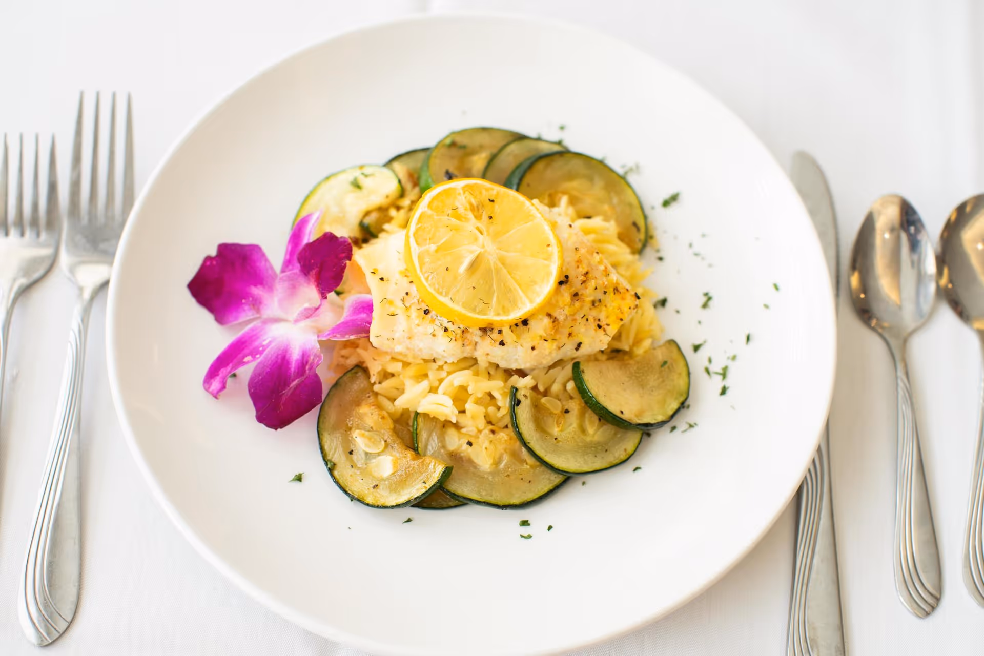 Plated fish on rice topped with a lemon slice, surrounded by zucchini slices and a purple flower garnish with utensils on a white tablecloth.
