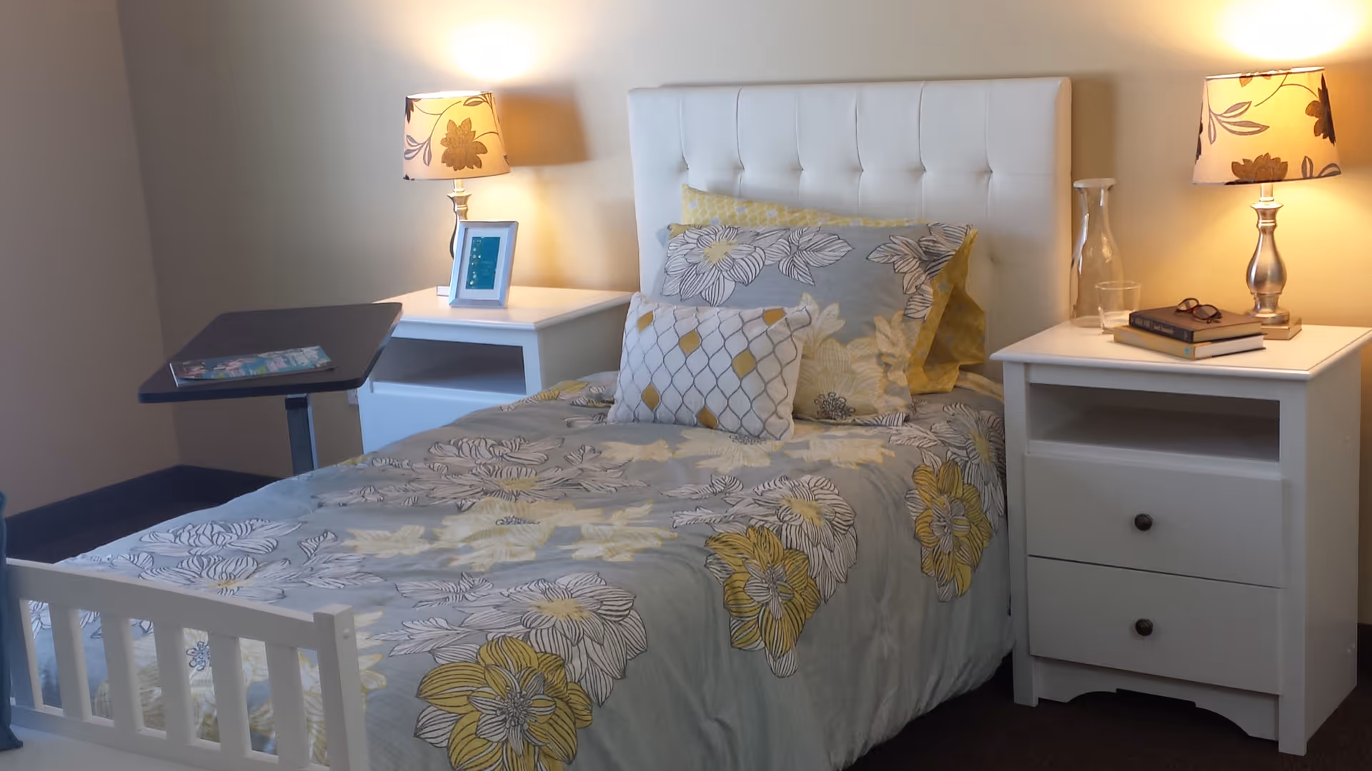 A neatly made single bedroom with floral bedding, a padded headboard, two nightstands with lamps, and an overbed table.