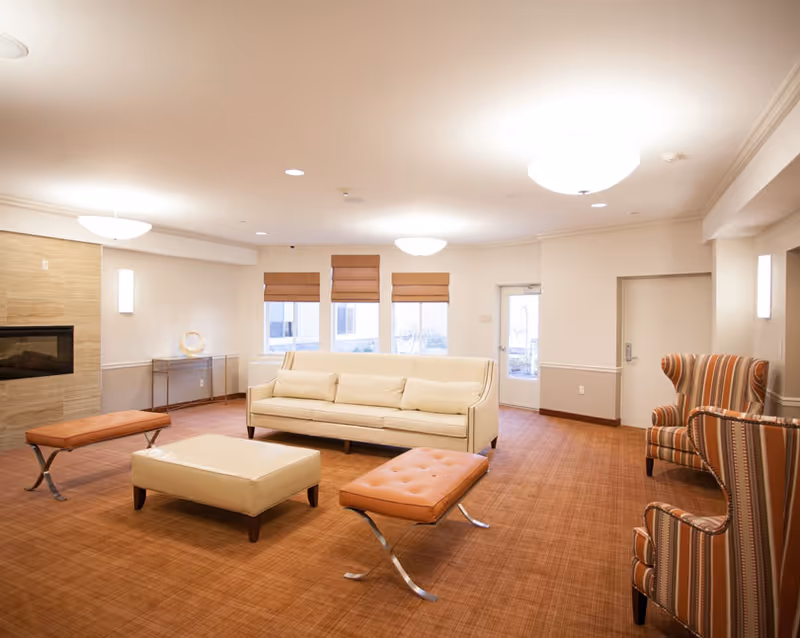 A bright and spacious living room with a beige sofa, two orange cushioned benches, a cream ottoman, and two striped armchairs. The room has a brown carpet, three windows with brown blinds, a modern fireplace on the left wall, and ceiling lights providing warm illumination.