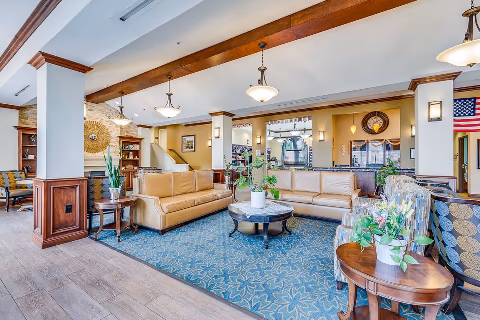 A spacious and well-lit senior living facility common area featuring two tan leather sofas arranged around a round coffee table with a potted plant. The room has a blue patterned rug, wooden beams on the ceiling, and multiple hanging light fixtures. There are additional chairs with patterned upholstery, wooden side tables with plants, and a stone fireplace with decorative elements. An American flag is visible in the background near a doorway.