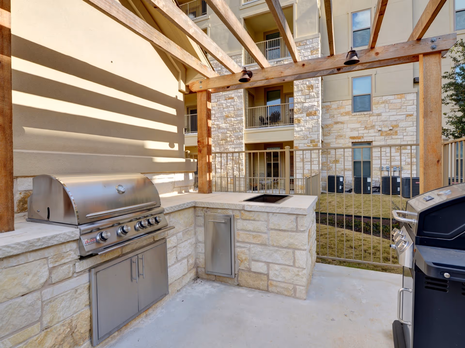 Outdoor grilling area with two stainless steel gas grills, stone countertops, and a wooden pergola overhead. The area is enclosed by a metal fence and is part of a multi-story building with balconies and windows visible in the background.