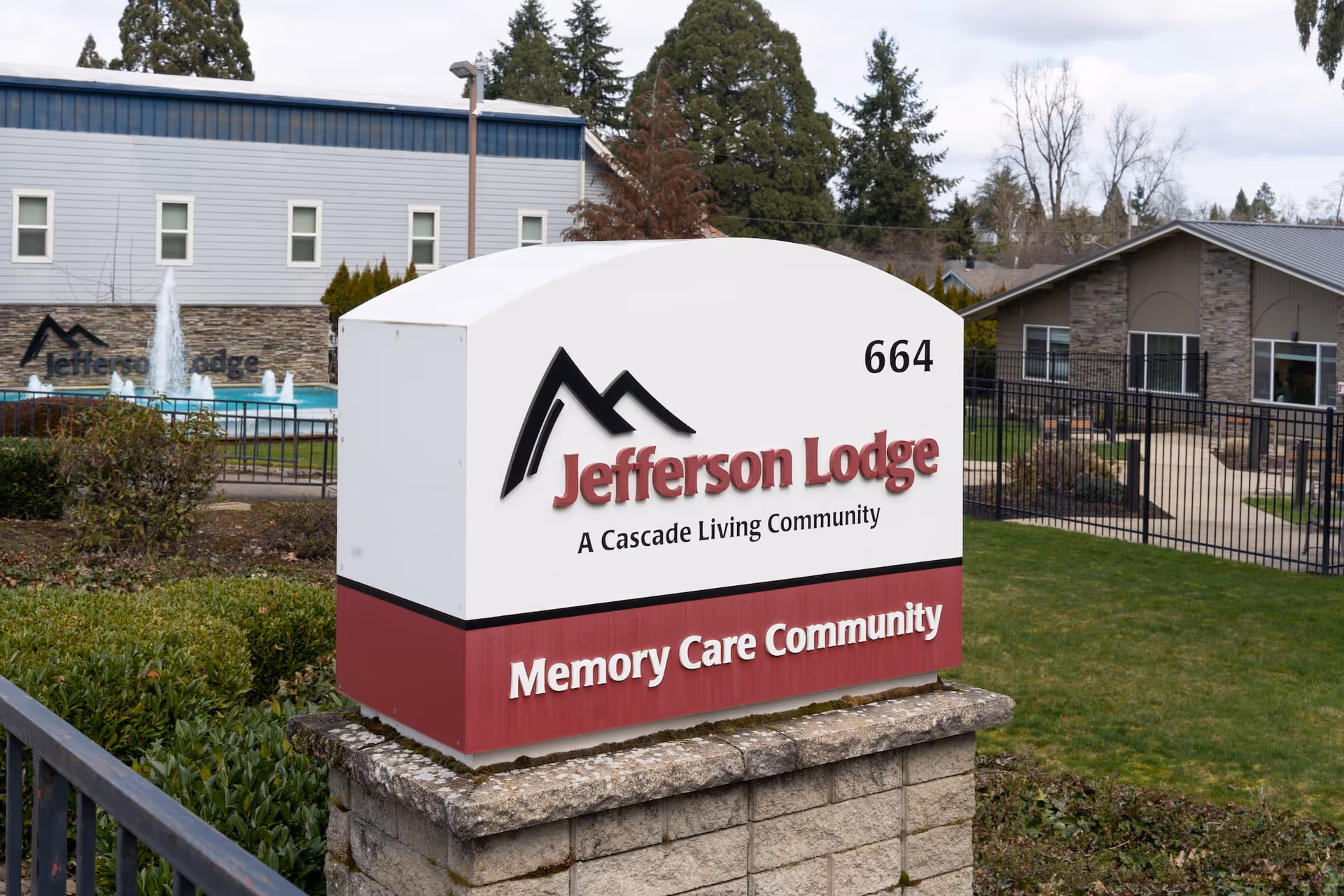Outdoor view of Jefferson Lodge Memory Care Community sign with buildings, a fountain, and greenery in the background.