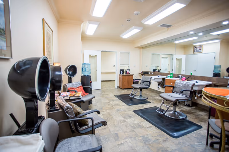 Interior salon area with hair dryers, styling chairs, wash sinks, and a large wall mirror in a senior living facility.