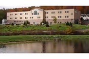 A large, three-story beige building with multiple windows, situated on a grassy hill with trees in the background showing autumn foliage. In the foreground, there is a pond reflecting the building and surrounding landscape.