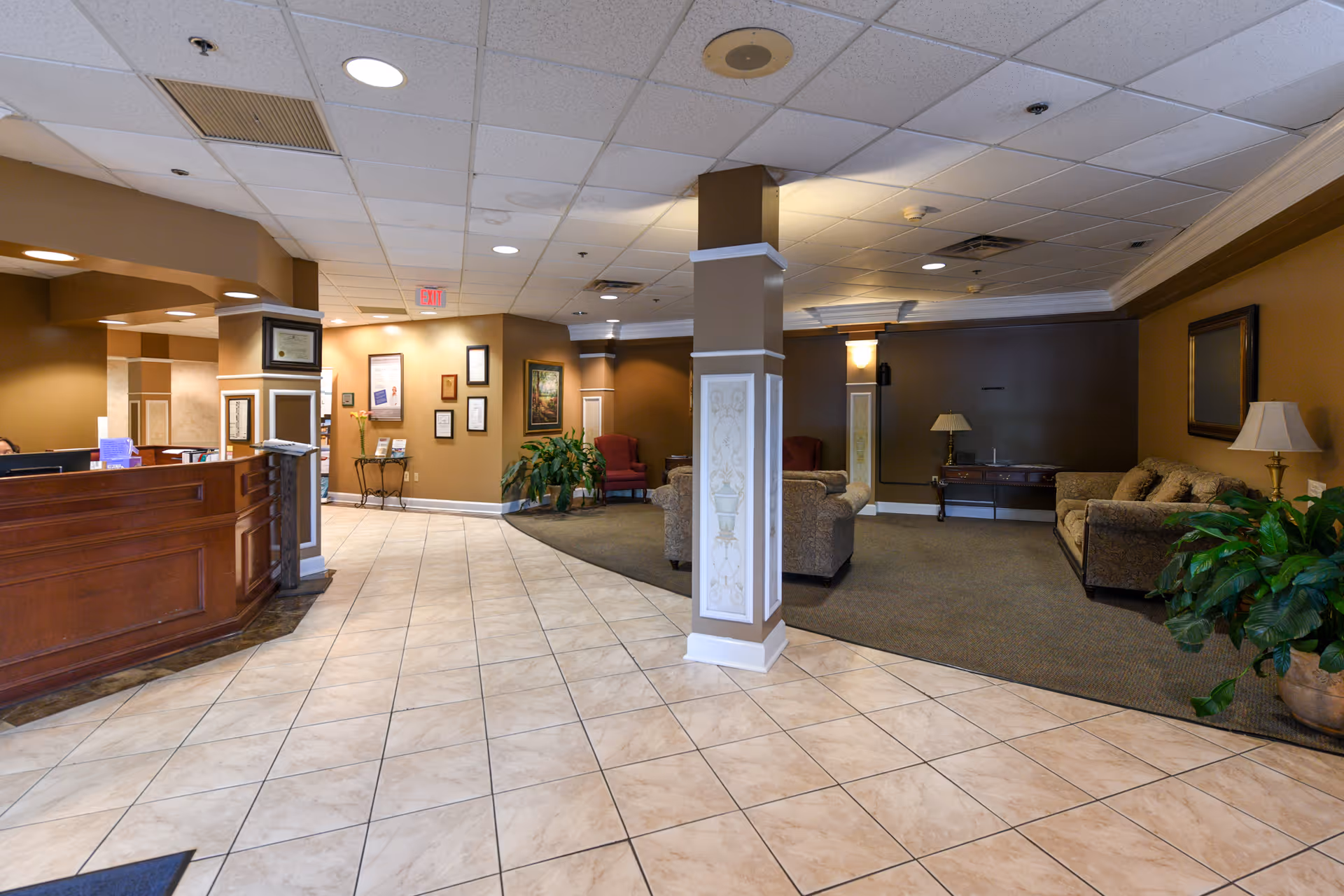 Reception lobby of a senior living facility with a wooden front desk, tiled floor, central decorative column, and a carpeted seating area.