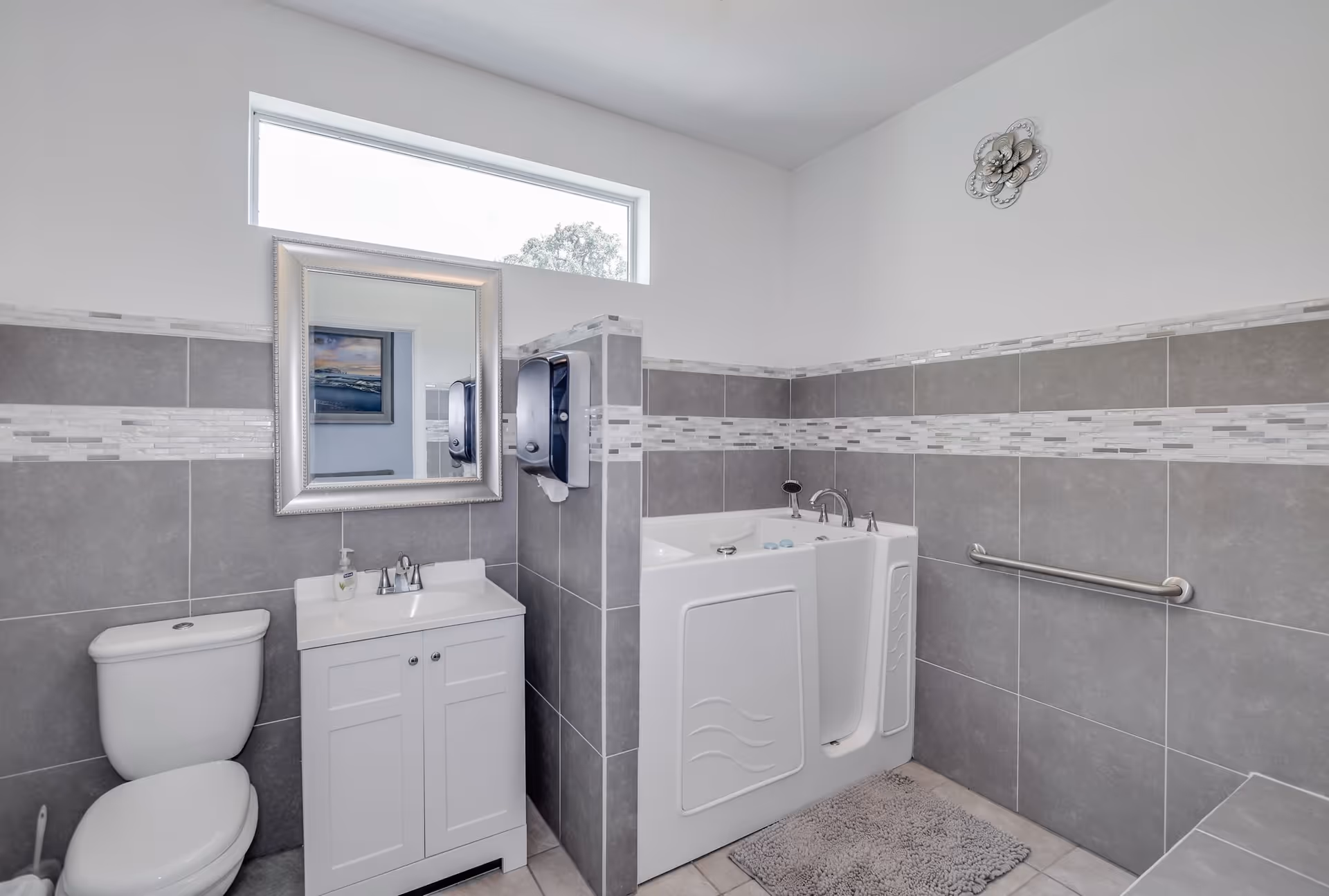 A modern bathroom with gray and white tiled walls, featuring a white toilet, a white vanity with a sink and a silver-framed mirror above it. There is a walk-in bathtub with a door and grab bar, a soap dispenser mounted on the wall, a small window near the ceiling, and a decorative wall piece above the bathtub.