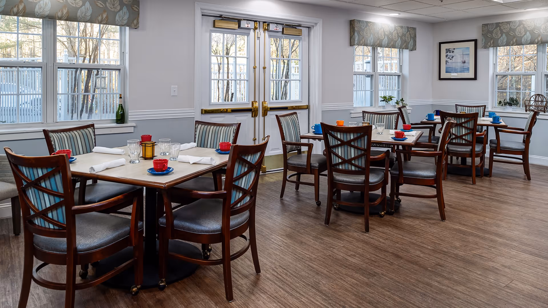 A bright dining room with several wooden tables and chairs arranged neatly. Each table is set with colorful cups, glasses, and white napkins. Large windows with patterned valances allow natural light to fill the room, and double doors with glass panes are visible in the background. The floor is wood, and the walls are painted light gray with white trim.
