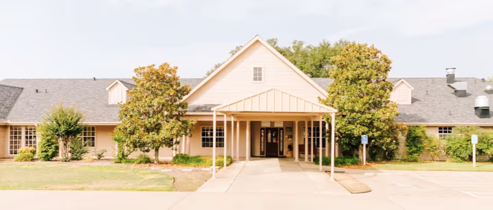 Front exterior view of a single-story senior living facility building with a covered entrance, surrounded by trees and greenery under a clear sky.