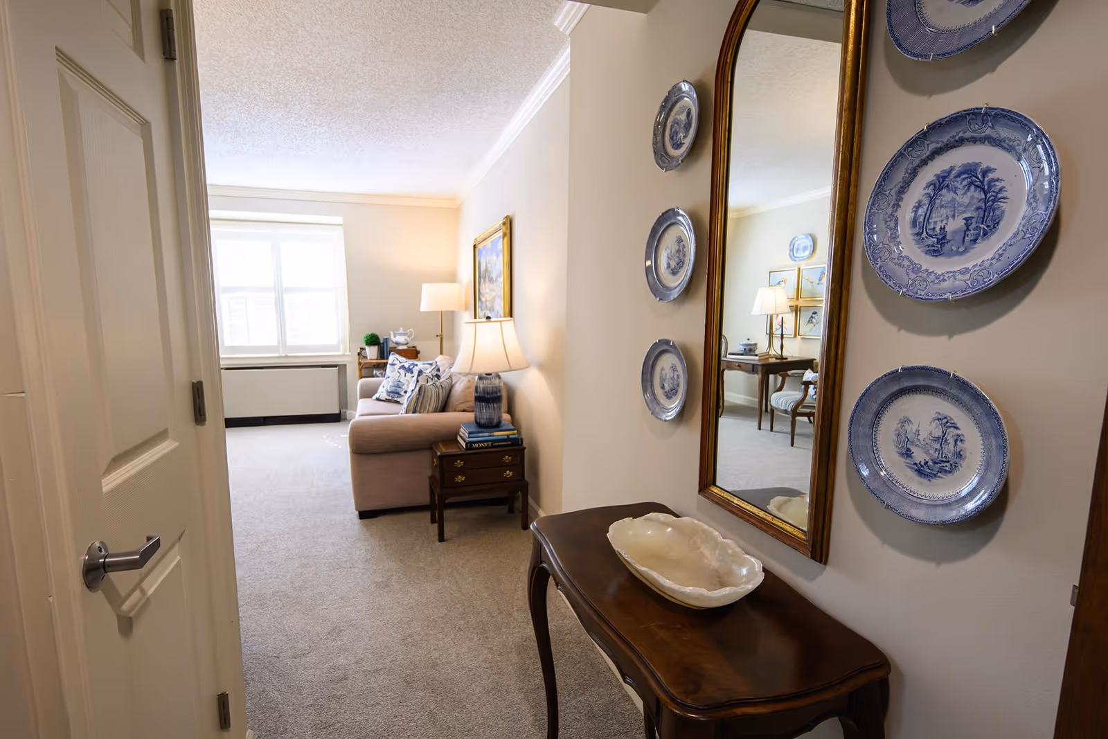 Entryway view into a cozy living room featuring a sofa, side tables with lamps, a console table with a decorative bowl, wall mirror and blue decorative plates.