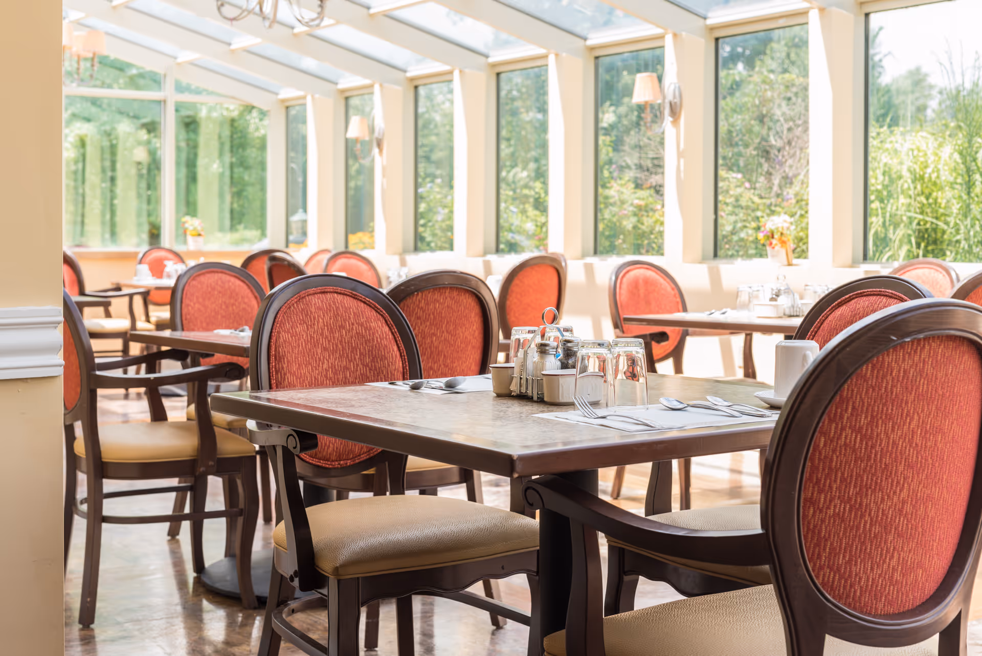 Sunlit dining room with wooden tables and red upholstered chairs set for meals by large windows.
