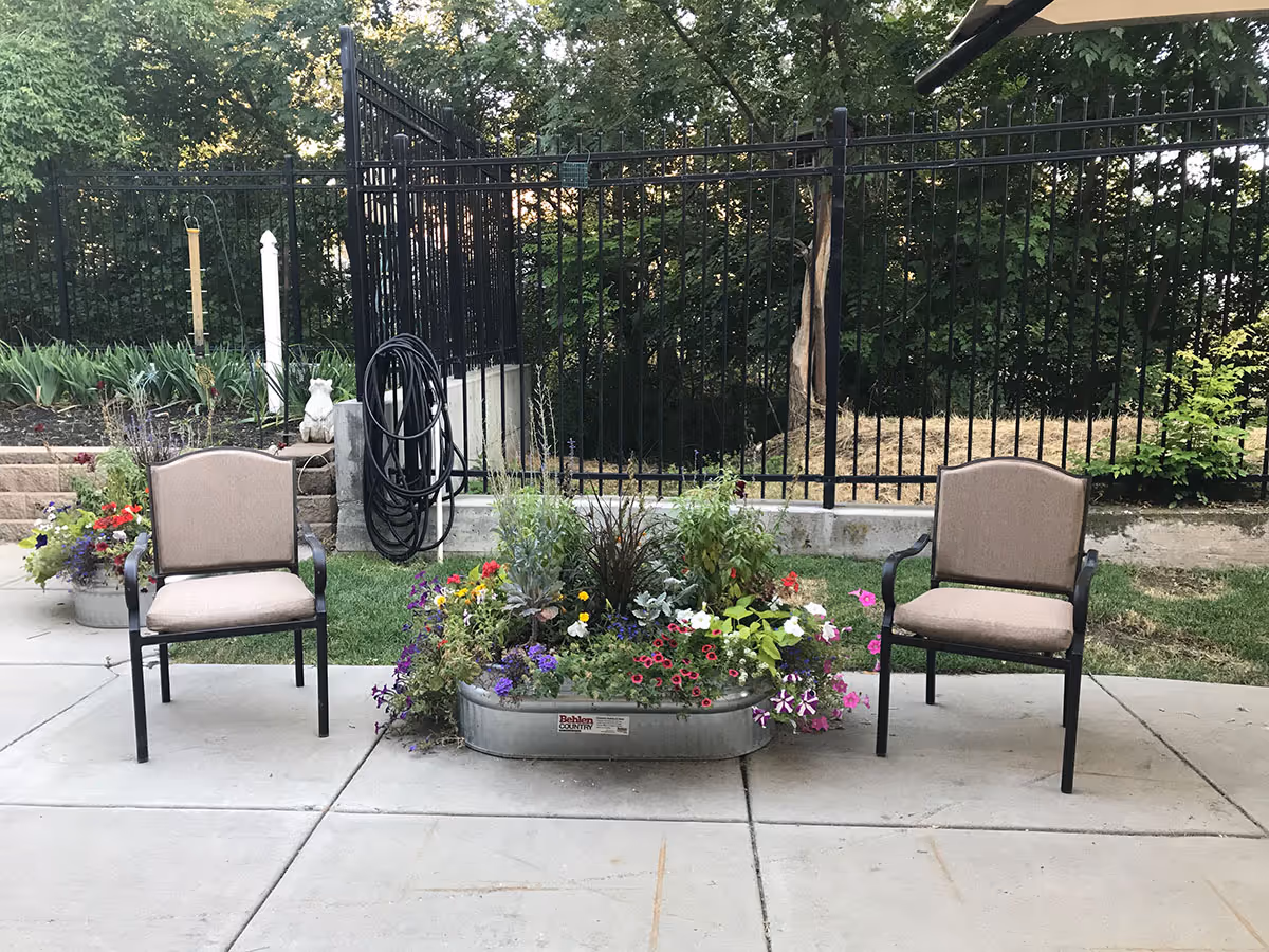 Two beige cushioned outdoor chairs placed on a concrete patio with a large metal planter filled with colorful flowers between them. Behind the chairs is a black metal fence with greenery and trees in the background. A coiled black hose hangs on the fence near the left chair.