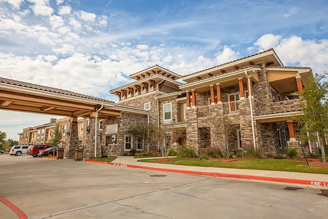Stone-clad assisted living building with a covered drop-off driveway and landscaping under a partly cloudy sky.