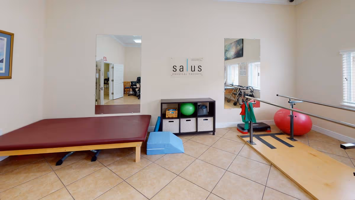 Physical therapy room with a treatment table, parallel bars, exercise ball, and shelving beneath a 'salus' sign.