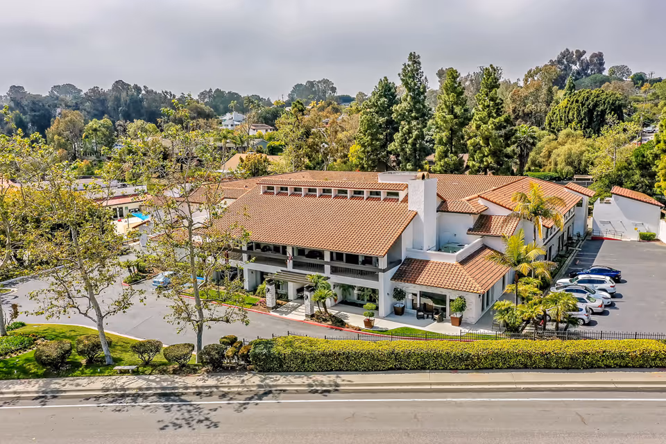 Aerial view of a two-story Mediterranean-style senior living facility with red tile roofs, parking lot and surrounding trees.